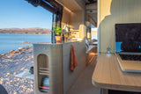 Modern RV rental interior with a laptop desk and kitchen, overlooking a serene lake and mountains. Perfect for van life!