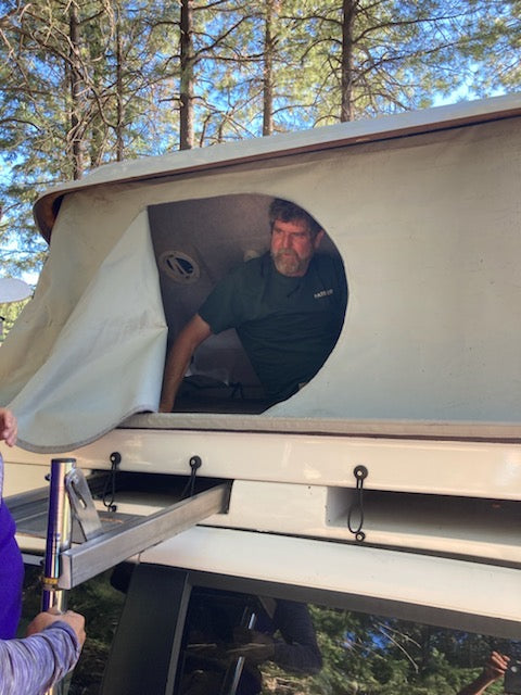 Bearded man emerges from a rooftop tent on an rv rental, with a ladder for access. Outdoor camping setup shown.