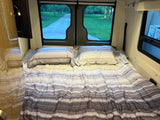 Cozy RV rental bed with striped bedding and a view of a park-like setting from the rear windows.