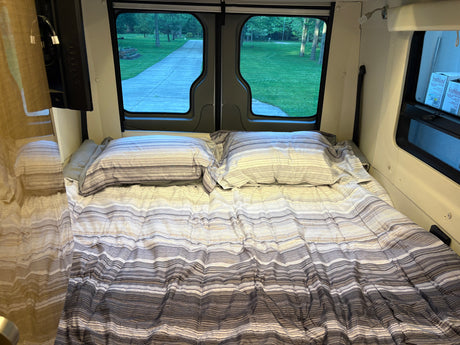 Cozy RV rental bed with striped bedding and a view of a park-like setting from the rear windows.
