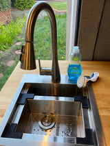 Compact RV rental kitchen sink with a modern faucet, Palmolive dish soap, and brush on a wood counter, overlooking greenery.