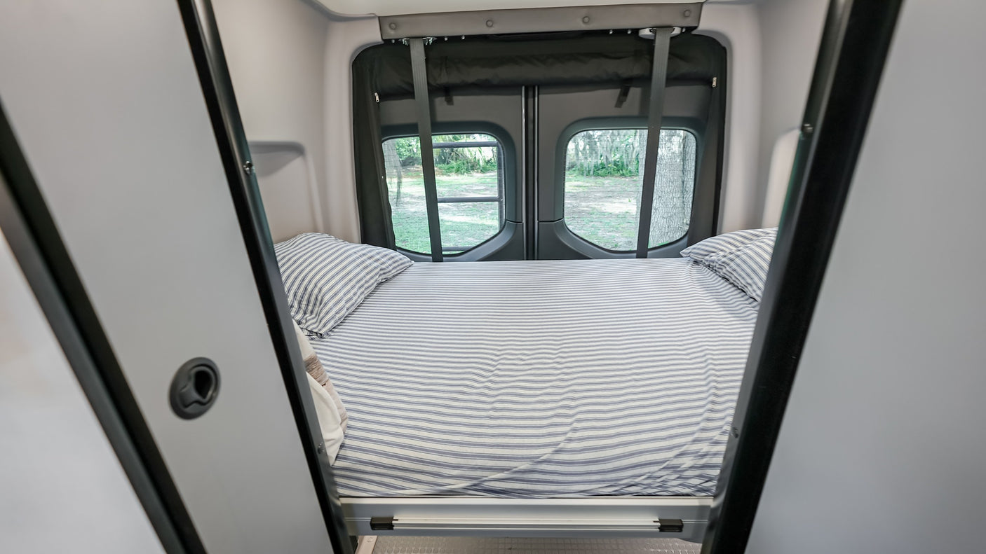 Neatly made bed in an rv rental, with striped sheets and green outdoor scenery visible through the rear windows.