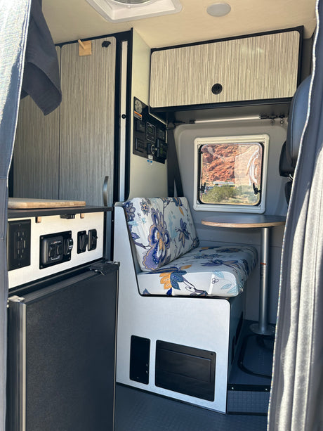 Cozy rv rental interior: floral dinette, window with desert view, kitchen counter with controls, and overhead storage.