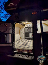 Inside a cozy teardrop RV rental: a comfy plaid bed awaits your next camping trip. Night view from outside.