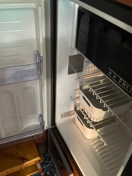 Open, empty compact RV rental refrigerator, revealing its interior with a freezer, wire shelves, and two storage bins.