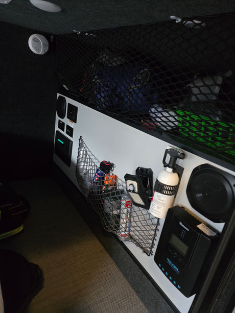 Explore a modern RV rental interior with organized cargo nets, control panel, Renogy solar charge controller, fire extinguisher, and detector.