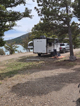 RV rental at a scenic campsite: a "GO LITE" travel trailer and truck by a calm lake, surrounded by pine trees and gravel ground.