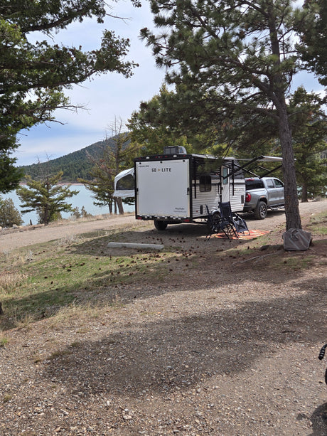 RV rental at a scenic campsite: a "GO LITE" travel trailer and truck by a calm lake, surrounded by pine trees and gravel ground.