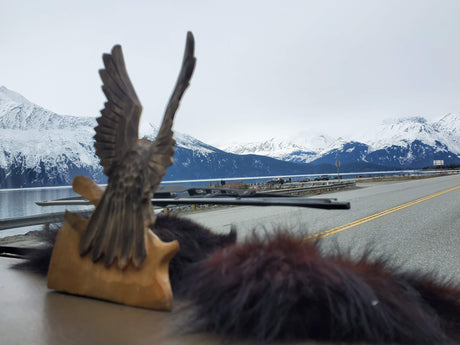 An RV rental journey: a carved bird statue on the dashboard faces a road, serene lake, and majestic snow-capped mountains.