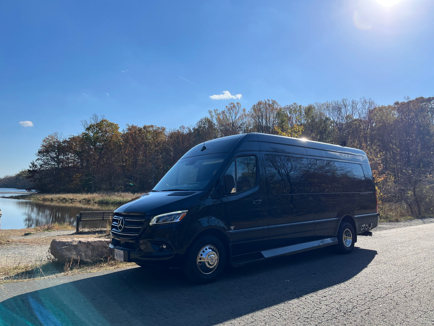 Black Mercedes-Benz Sprinter luxury van, an ideal rv rental, parked by a lake with autumn foliage.