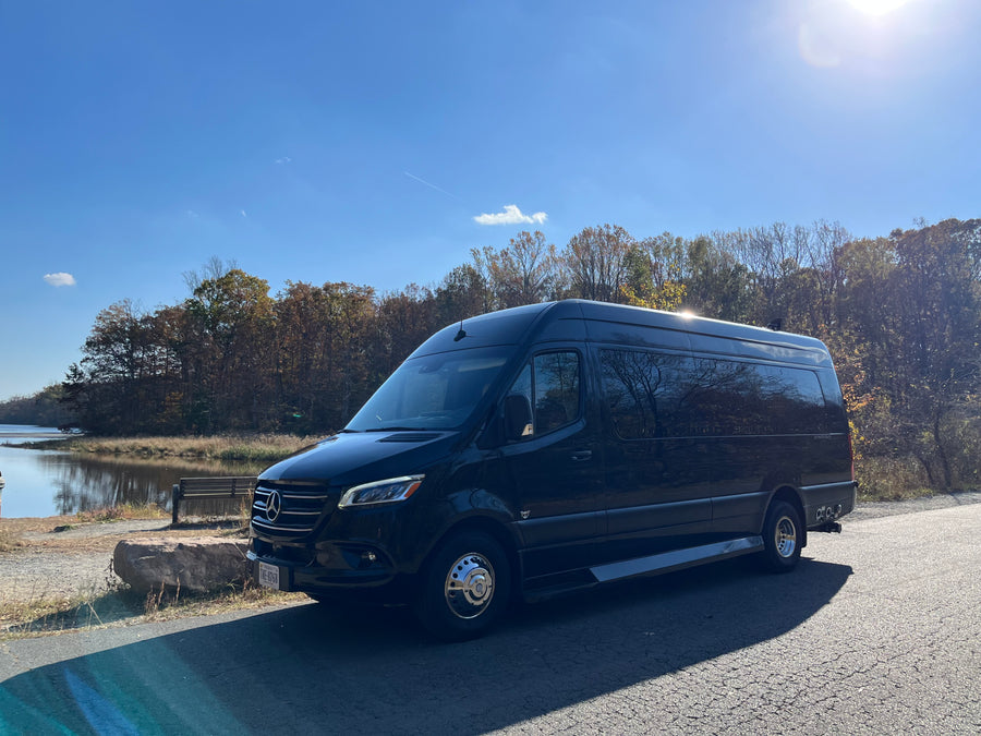 Black Mercedes-Benz Sprinter luxury van, an ideal rv rental, parked by a lake with autumn foliage.