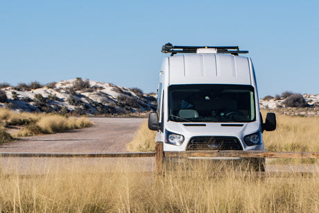 White RV rental camper van with rooftop equipment, parked behind a wooden fence in a sunny, dry landscape with sandy hills.