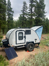 Enjoy an RV rental teardrop trailer with an extended awning and solar panel, parked in a serene forest campsite.