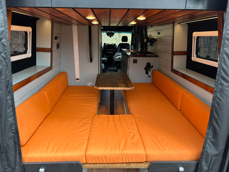 View of a camper van's inviting interior. Features include orange benches, a wooden table, and a compact kitchen. Great for rv rental.