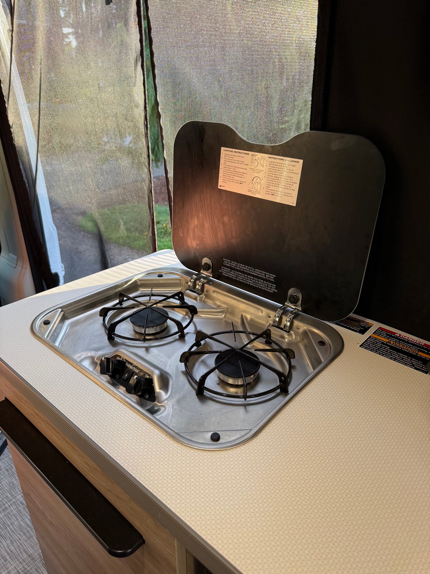 An RV rental kitchen featuring a two-burner gas stove with its lid open, revealing lighting instructions. A screened window shows greenery outside.