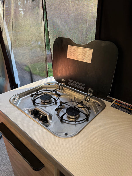 An RV rental kitchen featuring a two-burner gas stove with its lid open, revealing lighting instructions. A screened window shows greenery outside.