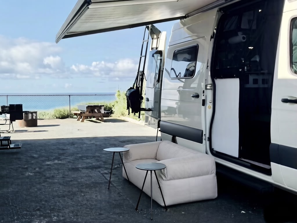 This RV rental offers an ocean view. A camper van with an awning, outdoor sofa, and tables is set at a scenic campsite.