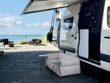 This RV rental offers an ocean view. A camper van with an awning, outdoor sofa, and tables is set at a scenic campsite.