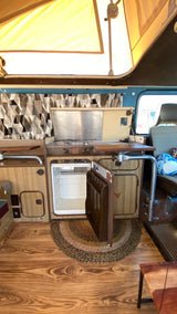 Inside a retro RV rental: a compact kitchen with a stove, sink, and open fridge, plus wood flooring and a pop-top.