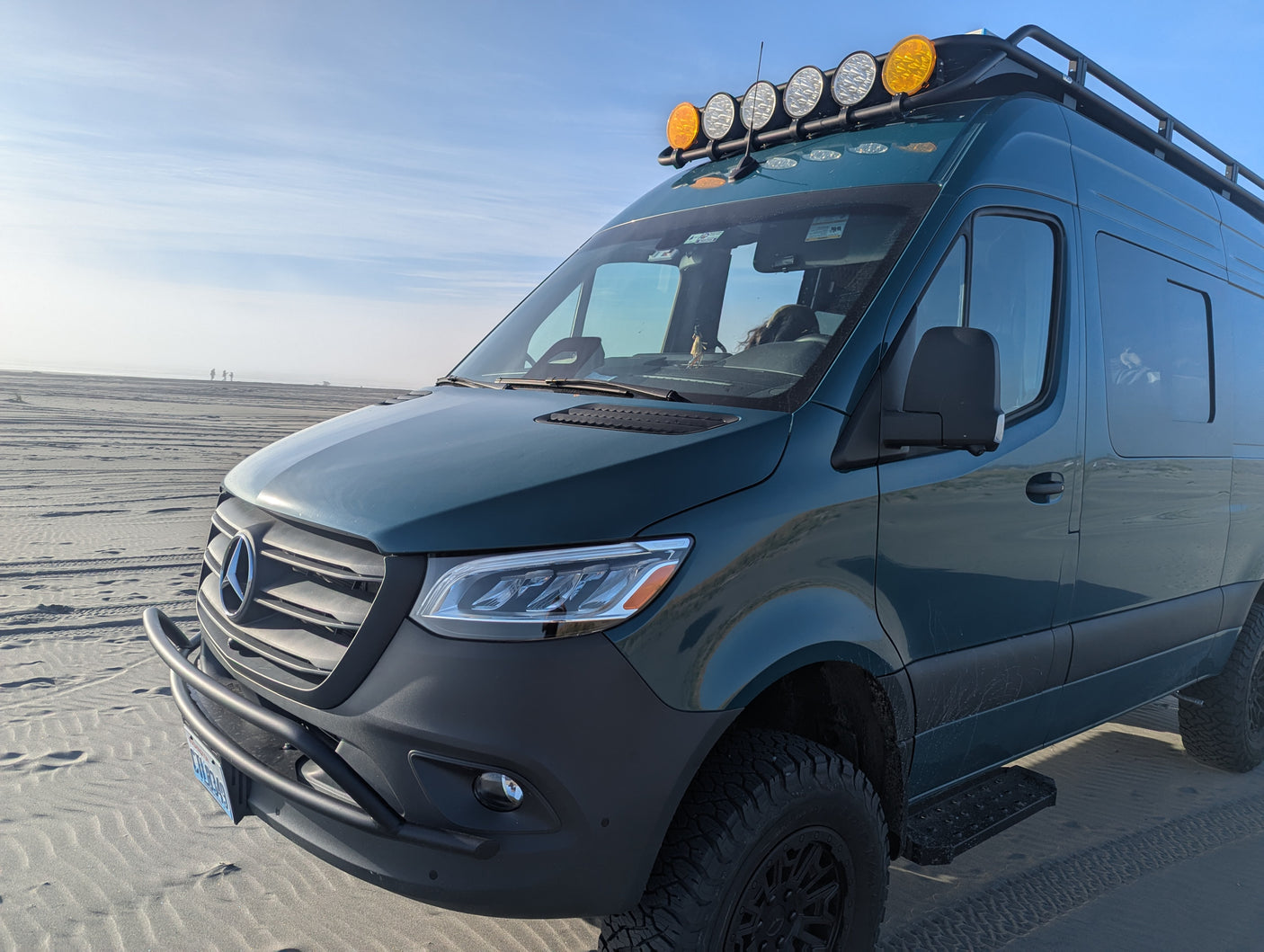 Explore with this custom Mercedes Sprinter rv rental, featuring off-road tires & roof lights, parked on a vast sandy beach.