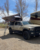 A tan Ram truck with an open rooftop tent & ladder sits on gravel next to a large RV. Great for outdoor camping or an RV rental adventure.
