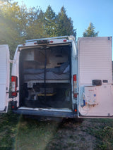 Open rear of an RV rental camper van revealing a cozy bed, storage, and an outdoor shower, set against trees & blue sky.