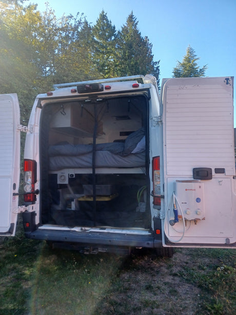 Open rear of an RV rental camper van revealing a cozy bed, storage, and an outdoor shower, set against trees & blue sky.