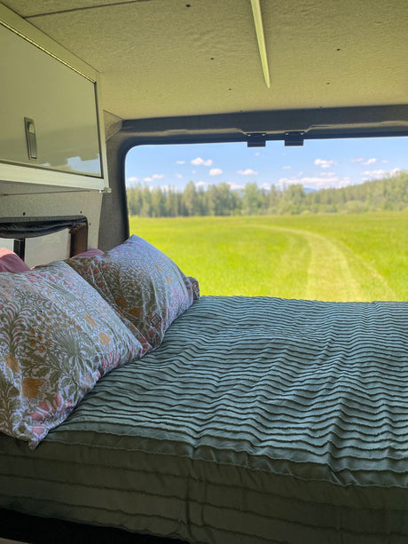 RV rental: interior bed with floral pillows, offering a window view of a vast, bright green field.
