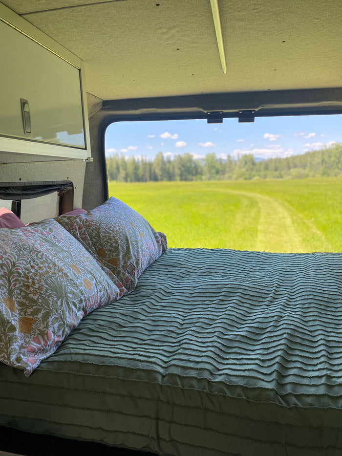 RV rental: interior bed with floral pillows, offering a window view of a vast, bright green field.