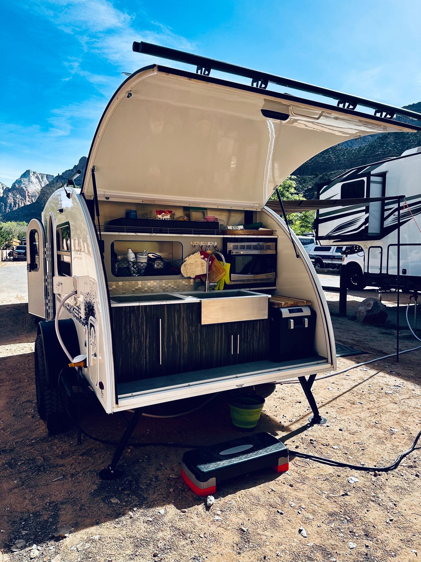  and picnic table make for a peaceful waterfront camping spot.|White teardrop RV rental showcasing its setup with a brown awning and patterned outdoor mat at a campsite.|Dark SUV with roof racks hitches to a white teardrop RV rental — photo 7