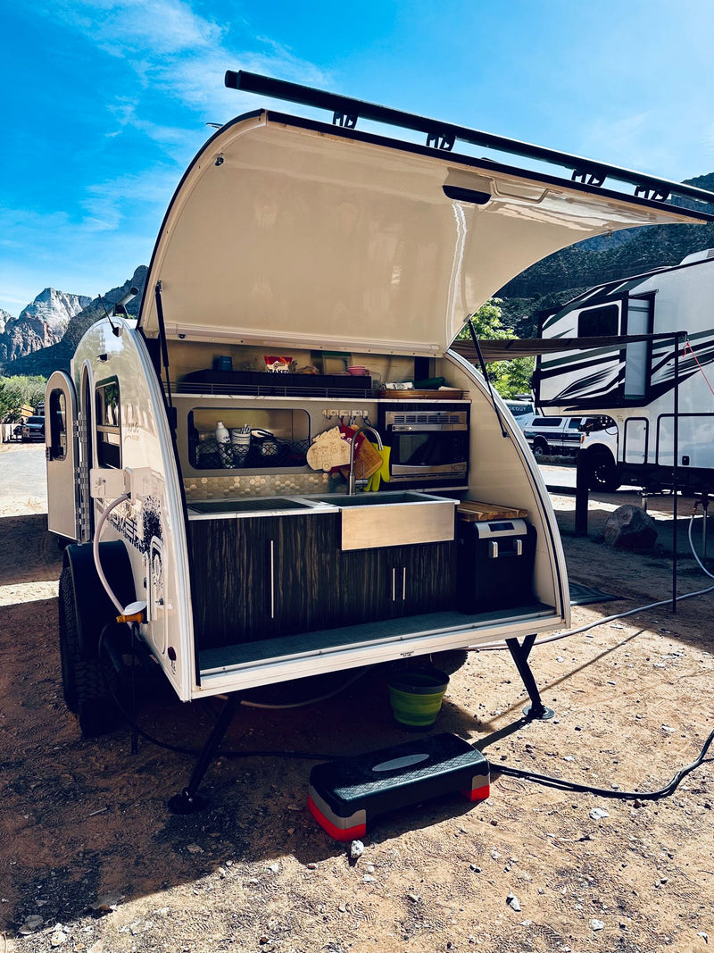  and picnic table make for a peaceful waterfront camping spot.|White teardrop RV rental showcasing its setup with a brown awning and patterned outdoor mat at a campsite.|Dark SUV with roof racks hitches to a white teardrop RV rental — photo 7