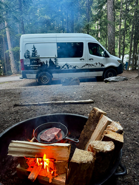 RV rental camping: Cooking steak on a campfire beside a WE adventure MT van amidst the trees.