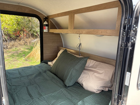 Inside an RV rental: cozy bed with view of nature, overhead storage, and fire extinguisher for safety and comfort.