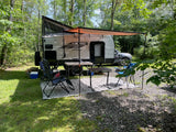 Viking pop-up RV rental comfortably set up at a wooded campsite, complete with awning, chairs, and tables for outdoor enjoyment.