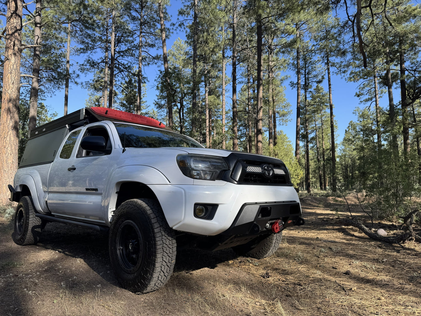 White Toyota Tacoma overland rig with a camper shell & red recovery boards in a sunny pine forest. Ideal for an rv rental adventure. — photo 10
