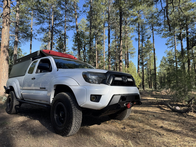 White Toyota Tacoma overland rig with a camper shell & red recovery boards in a sunny pine forest. Ideal for an rv rental adventure. — photo 10