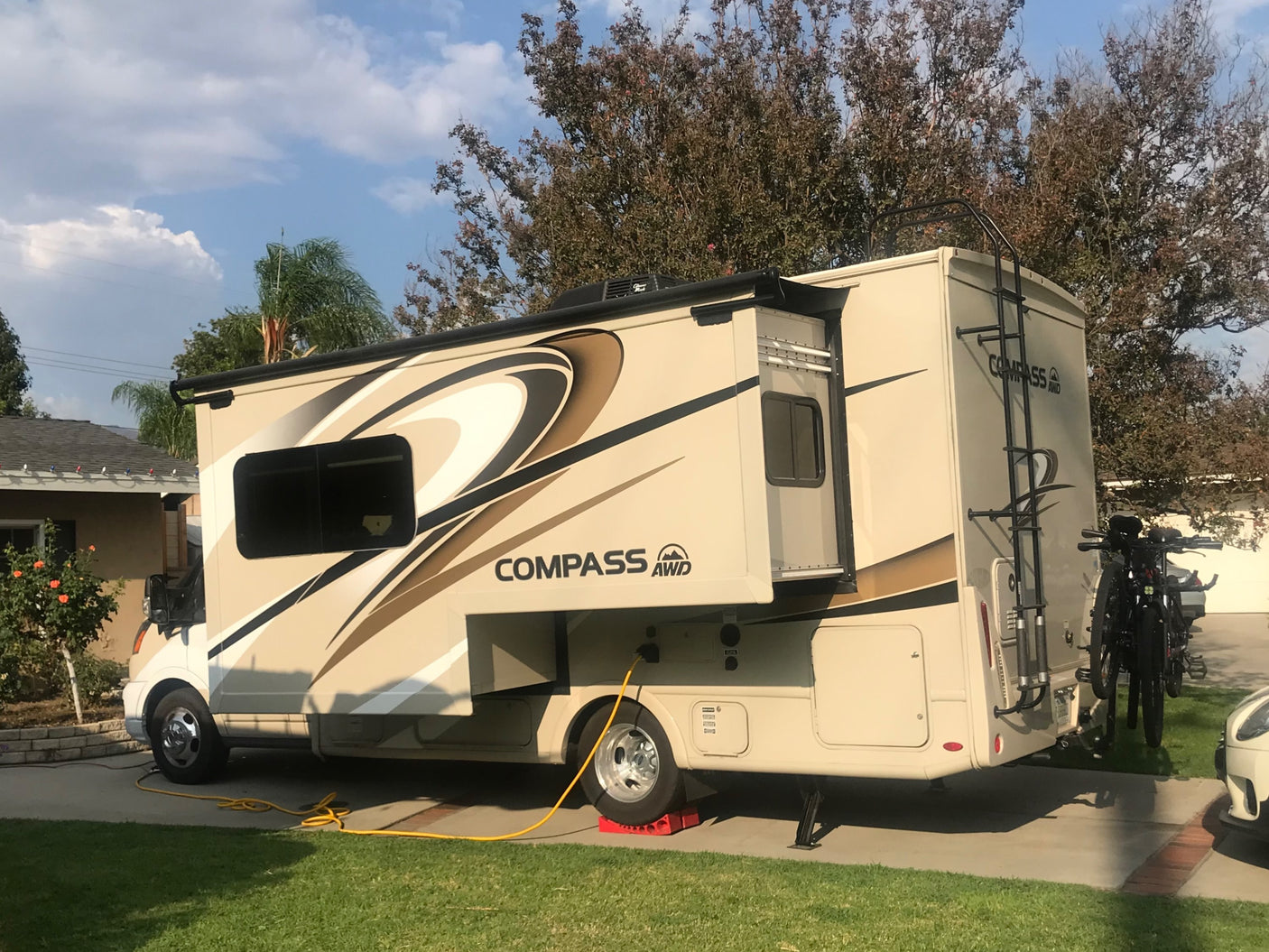 Beige RV rental, a Compass AWD model, parked on a driveway with an extended slide-out, bike rack, and power hookup.