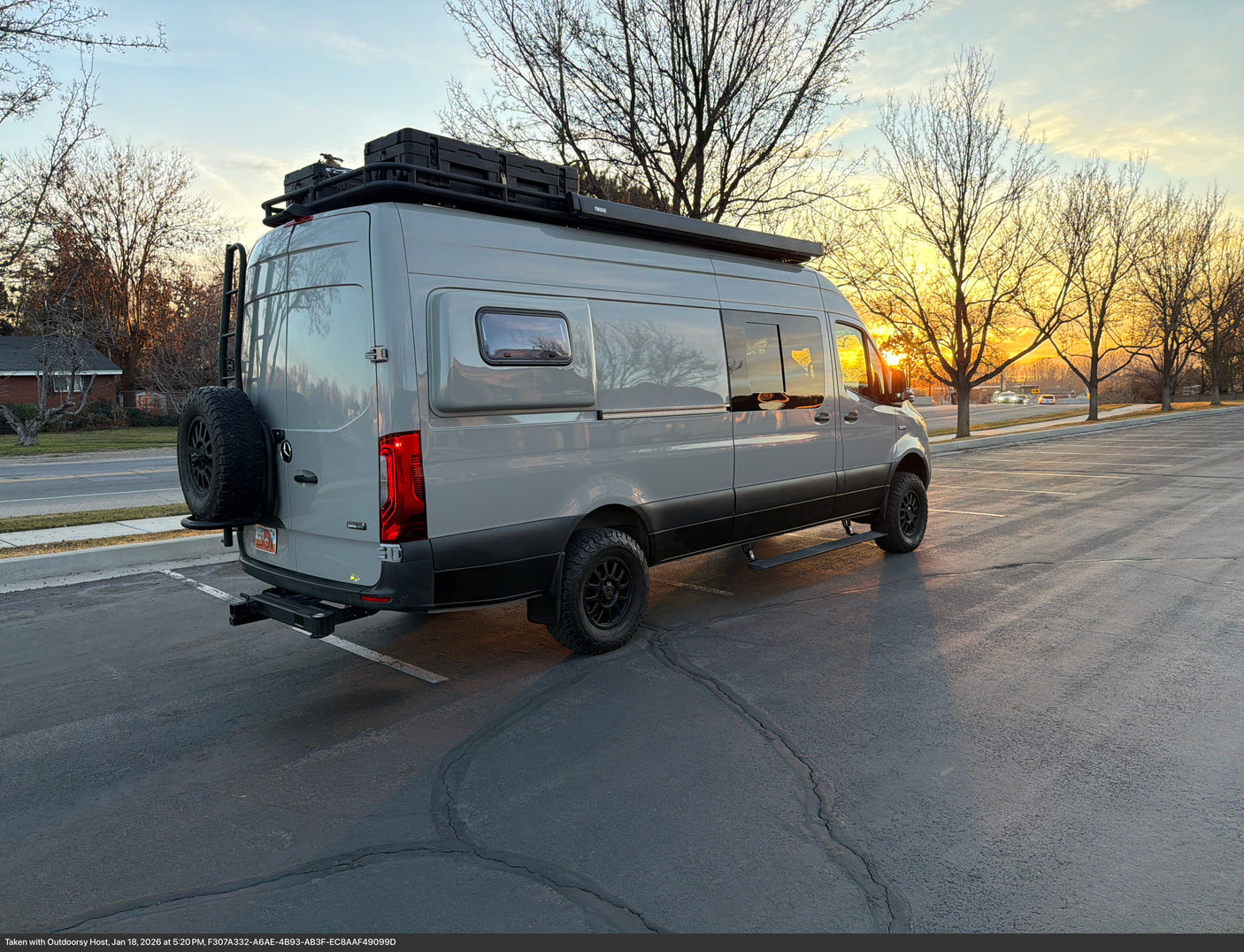 Gray Mercedes-Benz Sprinter camper van RV rental. Adventure-ready with roof rack, spare tire, and awning, seen at sunset.