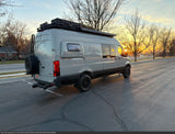 Gray Mercedes-Benz Sprinter camper van RV rental. Adventure-ready with roof rack, spare tire, and awning, seen at sunset.