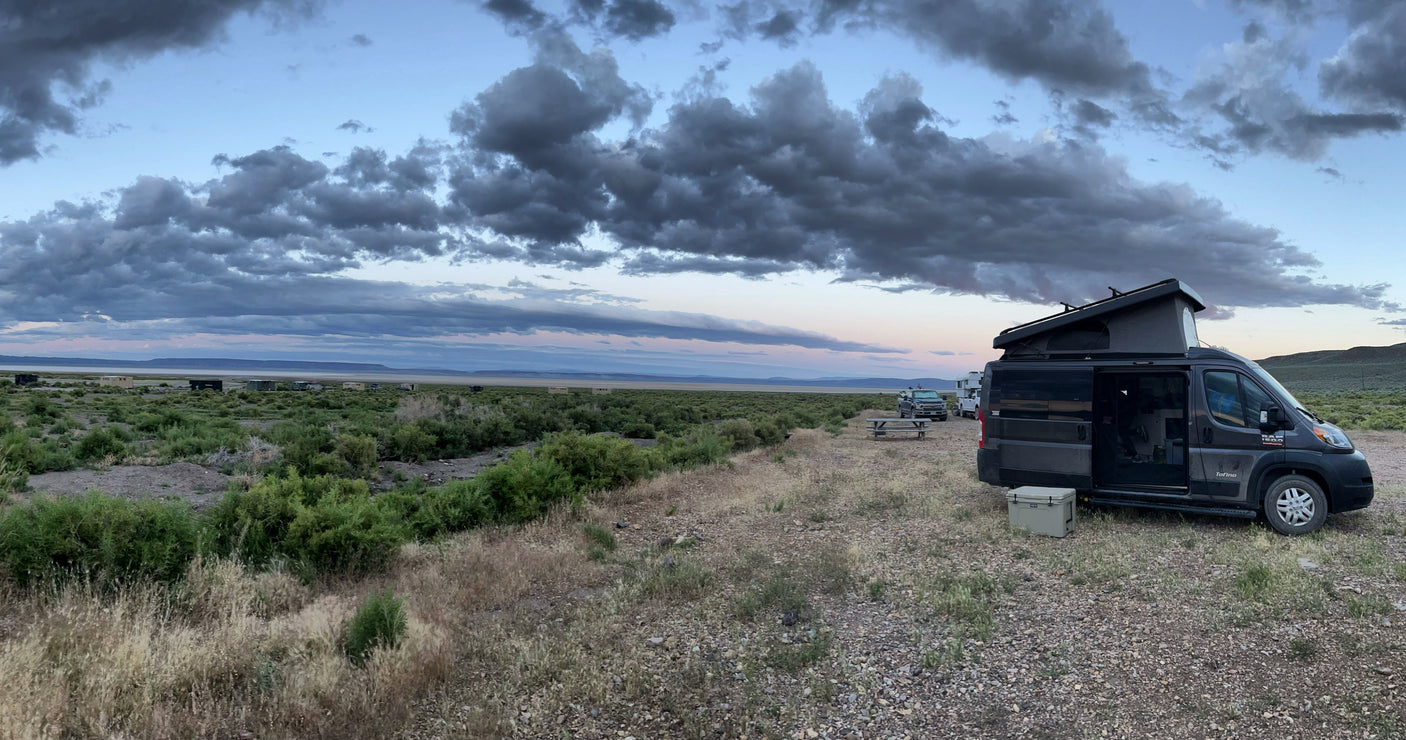 RV rental campervan set up for remote camping. A pop-top van sits on a gravel lot overlooking a vast, cloudy landscape at dusk.