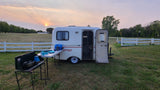 A small white RV rental is set up in a grassy field with an outdoor cooking station at sunset. Serene camping.