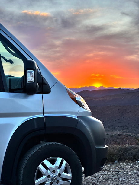 An RV rental camper van's front left side, parked on rocky ground, facing a vivid orange sunset over distant mountains.