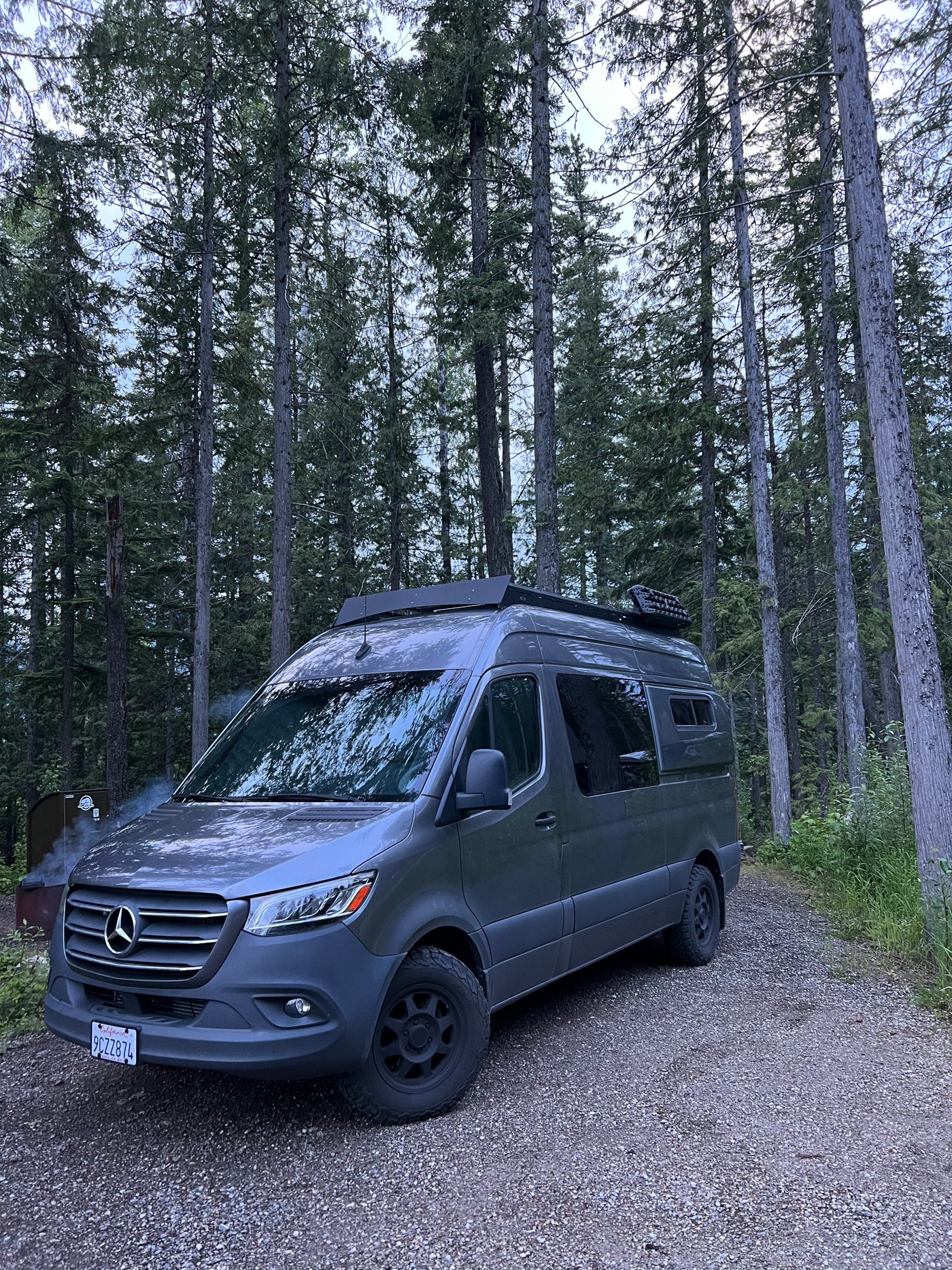 A grey Mercedes Sprinter camper van, an RV rental, is parked on a gravel road in a serene forest. Smoke rises from a nearby fire pit. — photo 3