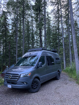 A grey Mercedes Sprinter camper van, an RV rental, is parked on a gravel road in a serene forest. Smoke rises from a nearby fire pit.