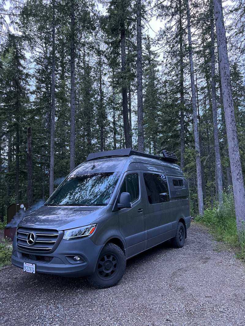 A grey Mercedes Sprinter camper van, an RV rental, is parked on a gravel road in a serene forest. Smoke rises from a nearby fire pit. — photo 3