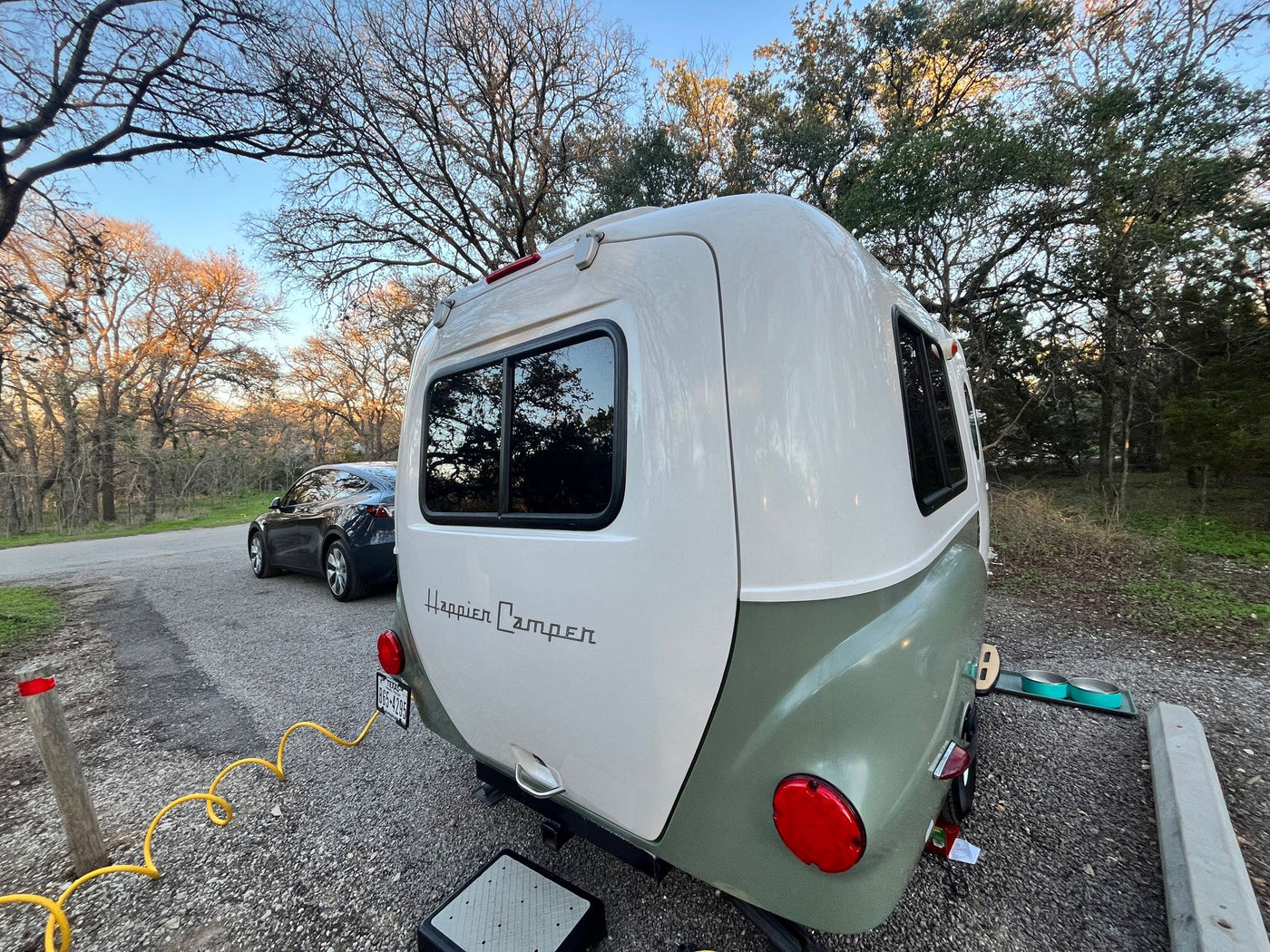 A two-tone Happier Camper HC1, ideal for an rv rental adventure, is set up at a gravel campsite with a car and pet bowls nearby. — photo 13