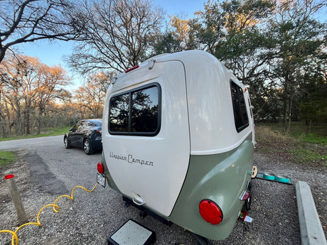 A two-tone Happier Camper HC1, ideal for an rv rental adventure, is set up at a gravel campsite with a car and pet bowls nearby.