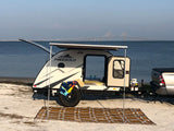 A Freesolo teardrop camper (RV rental) with awning extended on a sandy beach, overlooking a long bridge and calm water.