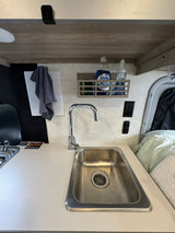 Inside an RV rental: a compact kitchen with a stainless steel sink, chrome faucet, wall-mounted shelf holding soap, and adjacent cooktop.