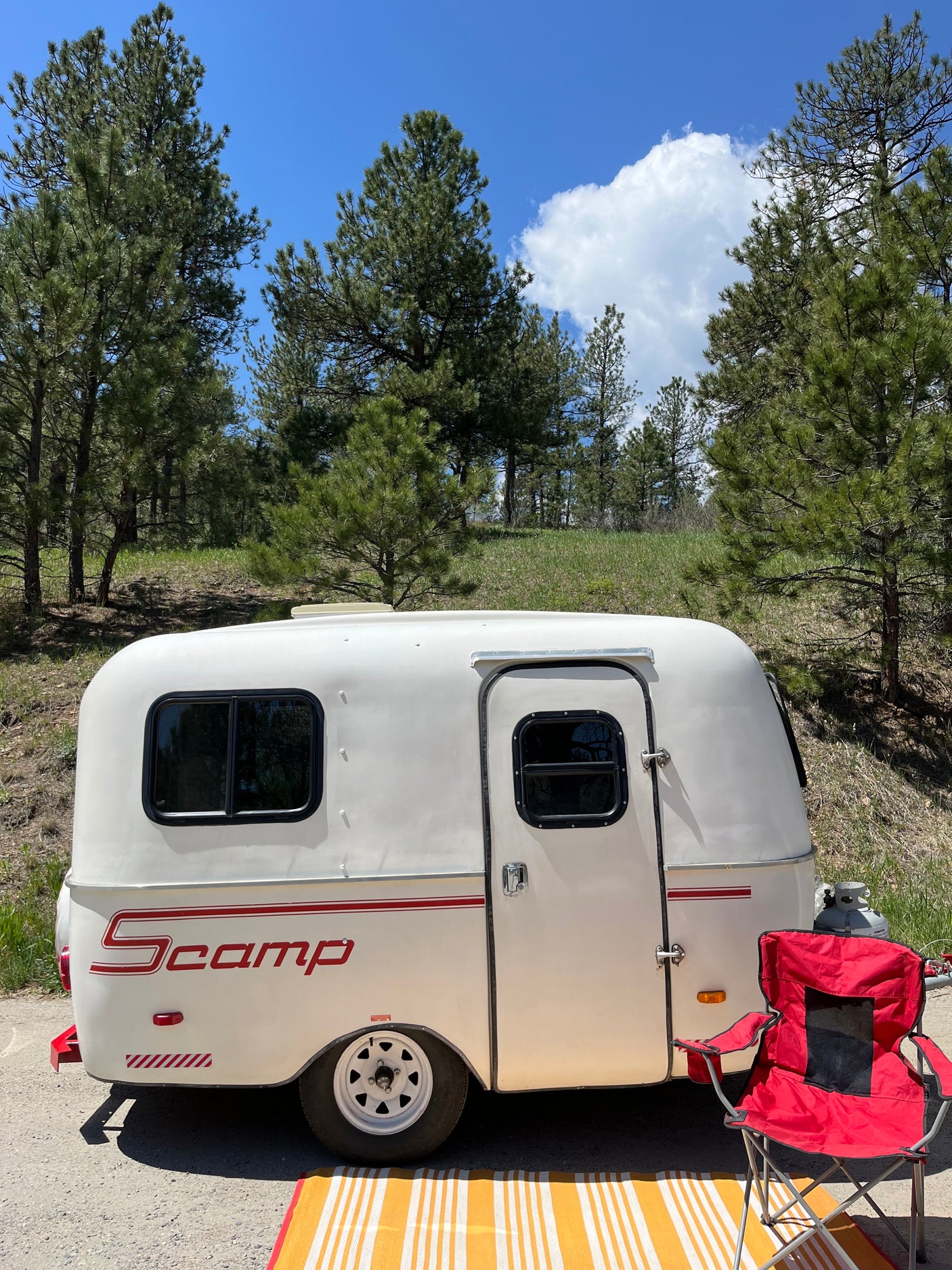 Enjoying nature with an RV rental: a white Scamp trailer by pine trees with a red chair and striped mat for outdoor relaxation.